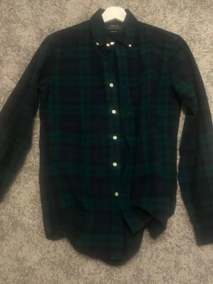 Rutig skjorta från Polo Ralph Lauren - Mörkgrön och marinblå rutig skjorta från Polo Ralph Lauren i custom fit. Skjortan har klassisk krage, långa ärmar och vita knappar framtill. Tillverkad i mjuk bomull, perfekt för en avslappnad men ändå stilren look.