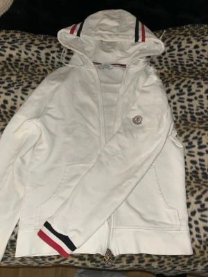Vit hoodie från Moncler - Vit hoodie från Moncler med dragkedja framtill, huva och fickor. Detaljer i rött, vitt och marinblått på huvan och ärmsluten samt Moncler-logga på bröstet. 