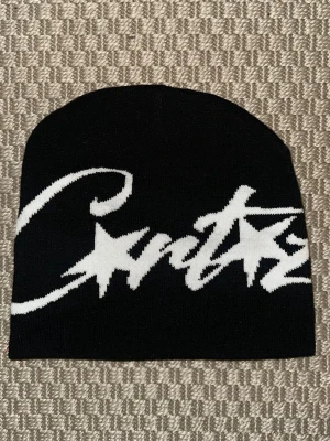 Corteiz mössa svart med vit logga - Säljer en svart mössa från Corteiz med stor vit logga och stjärndetaljer framtill. Mössan är ribbstickad och har en klassisk, tight passform. Perfekt för dig som gillar streetwear och vill sticka ut med en snygg accessoar.