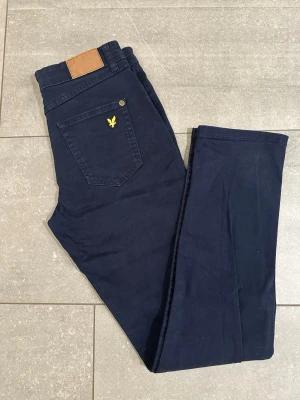 Mörkblå jeans från Lyle & Scott Junior - Snygga mörkblå jeans från Lyle & Scott Junior med klassisk femficksdesign och gul broderad logga på bakfickan. Jeansen har rak passform och är tillverkade i mjuk bomullsblandning för skön känsla. Perfekta för en avslappnad och stilren look.