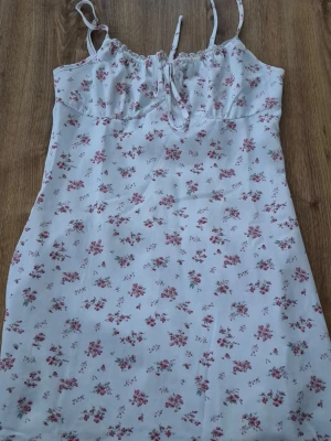 Vit blommig kortklänning med snörning - Söt vit kortklänning med små rosa blommor och tunna axelband. Klänningen har snörning vid bysten och är tillverkad i ett mjukt, lätt material som passar perfekt till varma dagar. Den har en feminin och avslappnad passform.