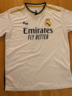 Real Madrid Bellingham matchtröja - Vit Real Madrid fotbollströja med svarta och guldiga detaljer. Framsidan har klubbmärke och Emirates Fly Better-tryck, baksidan har Bellingham och nummer 5. Perfekt för fotbollsfans som vill visa sitt stöd på läktaren eller planen.