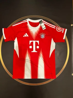 Bayern München matchtröja Adidas - Röd och vit Bayern München matchtröja från Adidas med klubbmärke och sponsorlogga på bröstet. Tröjan har vita detaljer, tre ränder på axlarna och texten 'FC Bayern München' på ryggen. Perfekt för fotbollsfans!
