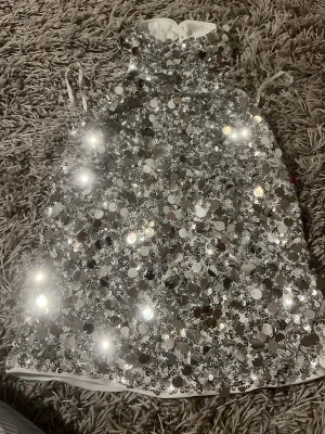 Glittrig silverpaljett topp - Supersnygg ärmlös topp täckt av stora och små silverpaljetter. Hög hals och öppen rygg ger en riktigt statement-look. Toppen  är fodrad i vitt tyg och har en loose passform. Perfekt för dig som vill sticka ut och glänsa.