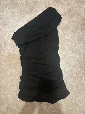 Svart oneshoulder kort klänning - Snygg svart tröja med oneshoulder-design. Klänningen har en åtsittande passform och är gjord i ett mjukt tyg som känns bekvämt mot huden. Perfekt för dig som vill ha en stilren och trendig look.