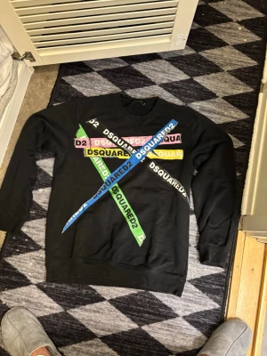 Svart DSQUARED2 sweatshirt med tryck - Cool svart sweatshirt från DSQUARED2 med färgglada logotexter i blått, grönt, rosa, vitt och gult som korsar varandra på framsidan. Tröjan har rund hals, långa ärmar och är gjord i mjukt bomullsmaterial. Perfekt för dig som gillar streetwear och statement-plagg.