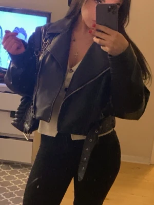 Svart biker skinnjacka med bälte - Snygg svart biker skinnjacka med breda slag och bälte i midjan. Jackan har dragkedja och en cool, lite oversized passform. Perfekt för dig som vill ha en edgy look till vardags. Passar till allt och ger en tuff vibe.