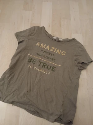 Grön t-shirt med text  - Beige t-shirt från H&M med glittrig text i guld och grönt: 'AMAZING FOREVER AND ALWAYS BE TRUE TO YOURSELF'. Klassisk rund halsringning och korta ärmar. Perfekt för en avslappnad stil.