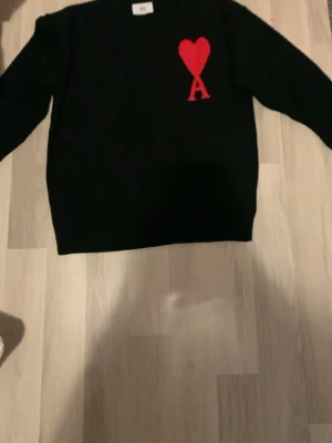 Svart stickad tröja från Ami Paris - Svart stickad tröja från Ami Paris med rött hjärta och bokstaven A broderat på bröstet. Klassisk rund halsringning och lång ärm. Perfekt för dig som gillar stilrena plagg med en twist.