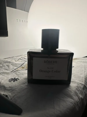 Nõberu Orange Cedar parfym - Upptäck Nõberu Orange Cedar No 109 Eau de Parfum i en stilren, mörk glasflaska med svart lock. Doften kombinerar fräscha toner av apelsin och cederträ för en unik och modern känsla. Flaskan rymmer 50 ml och har en minimalistisk etikett framtill. 40 available