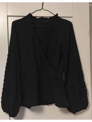 Svart omlottblus med volangdetaljer - Stilren svart omlottblus med volangdetaljer längs ringningen och långa puffärmar. Använd 1 gång💕