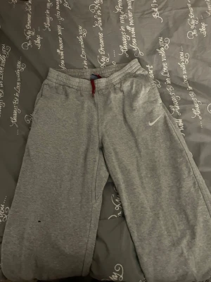 Grå mjukisbyxor från Nike  - Snygga grå mjukisbyxor från Nike i storlek XL med röd snörning i midjan, passar fortfarande mig som är storlek XS. och klassisk Nike-logga på benet. Byxorna har elastisk midja och är tillverkade i mjukt bomullsmaterial som är perfekt för chill eller träning.