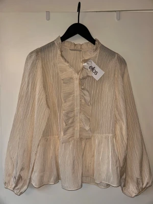 Krämvit volangblus från Ellos - Helt ny! Söt krämvit blus från Ellos med subtila ränder och volangdetaljer längs knäppningen. Blusen har långärmade puffärmar och lätt transparent tyg som ger en luftig känsla. Perfekt att styla med jeans eller kjol för en trendig look. Storlek 34/36 💕