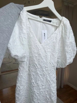 Vit kort klänning från Gina Tricot - Kort vit klänning från Gina Tricot med puffiga korta ärmar och v-ringning som är figursydd.  Klänningen har ett strukturerat, mönstrat tyg som ger en lyxig känsla. Perfekt för student.