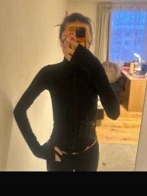 Svart  tröja med dragkedja - Säljer en svart, figurnära  tröja med dragkedja framtill och hög krage. Tröjan har långa ärmar och tumhål vid ärmsluten, vilket ger en sportig och modern känsla. Perfekt för lager-på-lager och passar till många olika stilar.