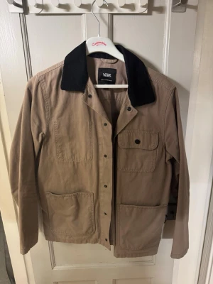 Beige overshirt från Vans - Stilren beige overshirt från Vans med svart krage i manchester. Jackan har tre stora fickor framtill och knappar längs hela framsidan. Perfekt att slänga över en hoodie eller t-shirt för en avslappnad look.