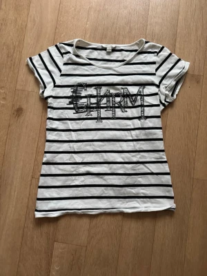 Randig t-shirt med CHARM-tryck - Vit och svart randig t-shirt från Bossini Ladies med texten 'CHARM' i paljetter på bröstet. Kortärmad modell med rund hals och mjukt bomullsmaterial. Perfekt för en avslappnad och trendig look.