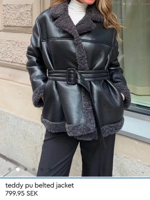 Svart teddyjacka med bälte Ginatricot - Snygg svart teddyjacka i PU-material med mjukt, mörkgrått teddyfoder och bred krage. Jackan har bälte i midjan, breda slag och långa ärmar med teddykanter. Perfekt för dig som vill ha en trendig och varm look.