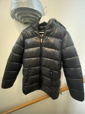 Svart dunjacka med huva - Snygg svart dunjacka med huva och dragkedja framtill. Jackan har två fickor med dragkedja och quiltad design som ger en klassisk pufferlook. Perfekt för kalla dagar och enkel att matcha med olika outfits.