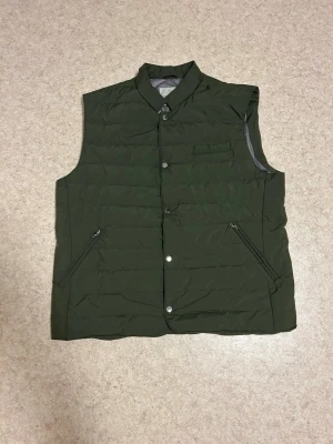 Grön vadderad väst från Four Hand Luxury strl M - Stilren och bekväm vadderad väst från Four Hands Luxury. Elegant mörkgrön färg som funkar perfekt både till vardag och mer uppklädda outfits. Västen har tryckknappar framtill samt två praktiska sidfickor med dragkedja, har även innerfickor på båda sidor.  Perfekt som lager-på-lager under höst och vår, eller över en stickad tröja för en klassisk look. Använd ett fåtal gånger men i skick som ny, inga skador eller fläckar.  Nypris ca 3000kr  📦 Skickas snabbt 🚭 Rök- och djurfritt hem