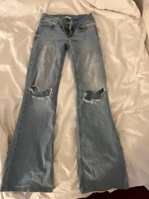Ljusblå bootcut jeans med hål - Säljer ett par ljusblå bootcut jeans med slitna hål över knäna och klassiska fem fickor. Jeansen har en avslappnad passform och utsvängda ben, perfekt för en trendig och avslappnad stil.