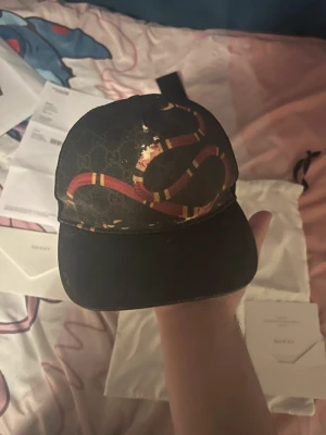 Gucci keps med ormtryck - Säljer en svart Gucci keps med det klassiska GG-mönstret och en stor röd och vit orm som slingrar sig över framsidan. Kepsen har böjd skärm och justerbar rem bak. Materialet är canvas med detaljer i läder. Perfekt för dig som vill sticka ut med en lyxig accessoar.