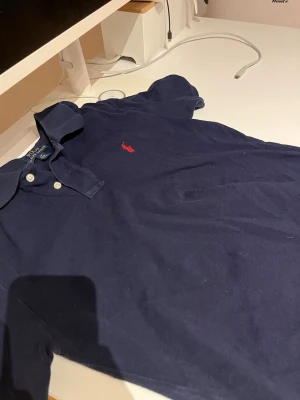 Marinblå pikétröja från Ralph Lauren - Klassisk marinblå pikétröja från Ralph Lauren med röd broderad logga på bröstet. Tröjan har krage, knappar vid halsen och är kortärmad. Perfekt för en stilren och avslappnad look.