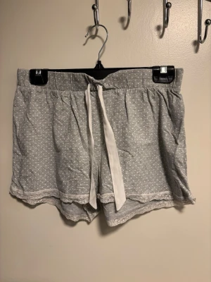 Grå pyjamasshorts med spetskant - Mysiga grå pyjamasshorts med små vita prickar och dekorativ spetskant längst ner. Shortsen har en bred resår i midjan och ett vitt knytband framtill för extra komfort. Perfekta för chillkvällar hemma eller sommarens varma nätter.