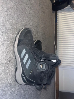 Svarta Adidas Terrex kängor - Säljer ett par svarta Adidas Terrex kängor med grå detaljer och klassiska tre ränder på sidan. Kängorna har snörning, robust sula och är tillverkade i slitstarkt syntetmaterial. Perfekta för utomhusaktiviteter och äventyr.