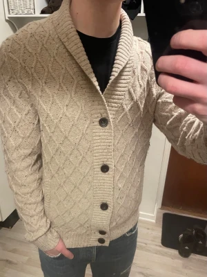 Beige stickad cardigan - Otroligt snygg beige stickad cardigan med mönster och bruna muddar vid armbågarna. Modellen är 180cm, 75kg. Priset kan såklart diskuteras!