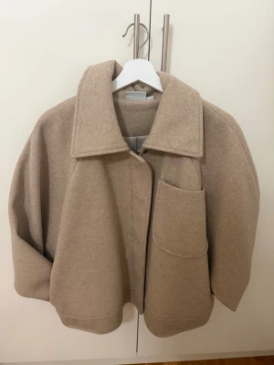 Beige oversized jacka - Säljer en beige oversized jacka med stor krage och unik stor ficka framtill. Jackan har en boxig passform och är tillverkad i ett tjockare ullmaterial som passar perfekt för kyligare dagar. Stängs med dold dragkedja framtill.