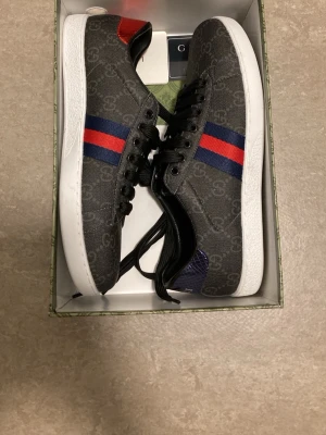 Gucci sneakers med GG-mönster - Snygga sneakers från Gucci med klassiskt GG-mönster i grått, vita sulor och svarta skosnören. Sidan har blå och röda ränder, samt detaljer i rött och blått på hälen. Perfekta för dig som vill sticka ut med stil.