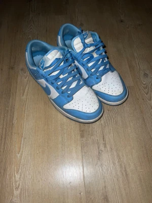 Nike Dunk Low blå och vita sneakers - Nike Dunk Low sneakers i blått och vitt med klassisk Swoosh-logga på sidan. Skorna har snörning, perforerad tå och platt sula. Ovandelen är i skinn och färgkombon ger en fräsch och sportig vibe. Perfekta för dig som gillar streetwear och vill sticka ut.