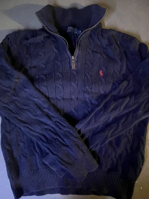 Mörkblå kabelstickad tröja Polo Ralph Lauren - Mörkblå kabelstickad tröja från Polo Ralph Lauren med halv dragkedja och röd broderad logga på bröstet. Tröjan har hög krage, långa ärmar och ribbade muddar. Perfekt för dig som gillar klassisk och tidlös stil. passar 170-180