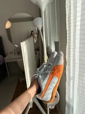 Puma Speedcat sneakers i grått och orange - Snygga Puma sneakers med ljusblå mocka och orangea detaljer längs sidan. Skorna har klassisk snörning, rund tå och en gummisula i naturfärg. Perfekta för dig som vill ha en sportig och trendig look.