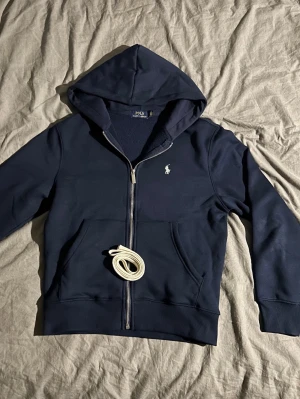 Marinblå hoodie från Polo Ralph Lauren - Snygg marinblå hoodie från Polo Ralph Lauren med dragkedja, huva och klassisk vit logga på bröstet. Mjuk insida och två stora fickor framtill. Perfekt för en chill och stilren look.