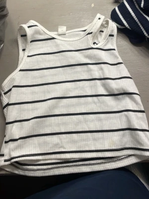 Vit ribbad crop top med svarta ränder - Säljer en vit ribbad crop top med svarta horisontella ränder. Toppen är ärmlös med breda axelband och har en avslappnad passform. Perfekt till sommaren och enkel att matcha med jeans eller kjol.