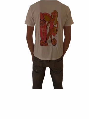Vit t-shirt med Beach Babe-tryck - Säljer en vit t-shirt i bomull med färgglatt Beach Babe-tryck på ryggen. Trycket har motiv av en tjej med solglasögon, surfbräda och blommor i rosa, orange och gul. Klassisk rund hals och korta ärmar. Perfekt för dig som gillar sommarvibbar och coola prints.