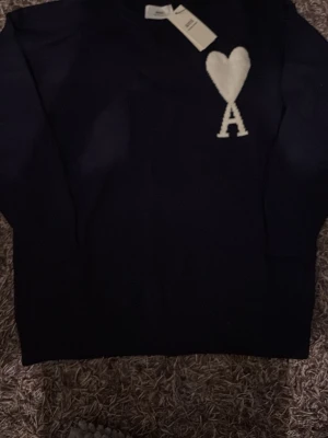 Svart stickad tröja från Ami Paris - Mörkblå stickad tröja från Ami Paris med deras ikoniska vita hjärta och bokstaven A broderat på bröstet. Klassisk rund halsringning och långärmad modell. Perfekt för en stilren och trendig look.