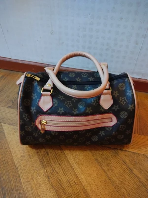 Svart handväska med mönster och dragkedja - Snygg svart handväska med beige handtag och detaljer. Väskan har ett klassiskt mönster med stjärnor och blommor, samt en praktisk dragkedja framtill. Materialet är skinnimitation och väskan har guldfärgade metalldetaljer. Perfekt storlek för dina viktigaste grejer.