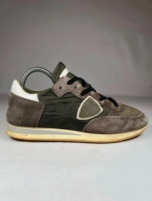 Philippe Model sneakers i olivgrönt - Snygga sneakers från Philippe Model i olivgrönt och beige med detaljer i mocka och mesh. Skorna har svarta snören, rund tå och en platt sula. Ikonisk sköldlogga på sidan och kontrastdetaljer i vitt. Perfekt för dig som gillar en avslappnad men stilren look.