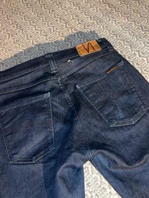 Mörkblå jeans från Nudie Jeans, W30 L30 - Säljer ett par mörkblå jeans från Nudie Jeans i klassisk rak modell. Jeansen har snygga bakfickor med Nudie-logga och läderpatch i midjan. Tillverkade i slitstarkt denimtyg, skriv för mer info 
