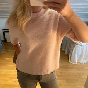 Ljusrosa stickad tröja med kort ärm - Supermjuk och fluffig ljusrosa stickad tröja med korta ärmar och rund hals. Tröjan har en avslappnad passform och ribbade kanter vid ärmslut och nederkant. Perfekt för lager-på-lager och ger en söt vibe till din outfit.