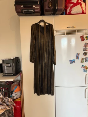 Glittrig långklänning från Cream - Snygg glittrig långklänning från Cream i guldigt material med v-ringning och långa ärmar. Klänningen har ett tunt, skimrande tyg och ett band vid halsen som kan knytas. Perfekt för dig som vill sticka ut med en cool och elegant look. Storlek L. Mycket fint skick, jag hittar inga synliga anmärkningar eller defekter. 