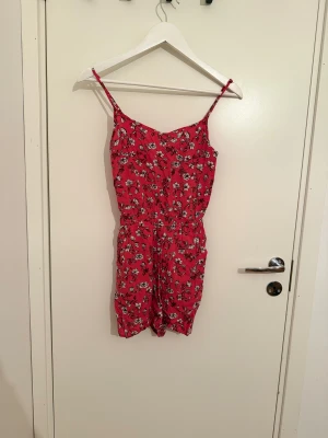 Röd blommig byxdress från H&M, strl 34 - Söt röd byxdress med vita och svarta blommor från H&M. Smala axelband, resår i midjan och korta ben. Perfekt för varma dagar och enkel att styla med sneakers eller sandaler.