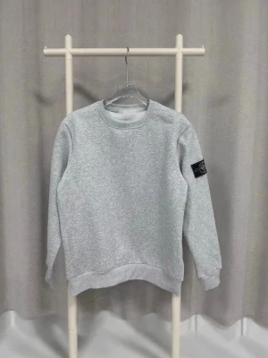 Grå sweatshirt från Stone Island - Snygg grå sweatshirt från Stone Island med klassisk rund hals och ribbade muddar. Tröjan har den ikoniska Stone Island-loggan på vänster ärm, fäst med två knappar. Perfekt för dig som gillar stilrena och sportiga plagg.