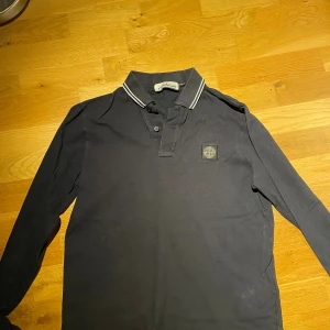 Svart långärmad piké från Stone Island - Svart långärmad pikétröja från Stone Island med klassisk krage och vita ränder på kragen. Tröjan har knappslå framtill och ikonisk logopatch på bröstet. Skön passform i bomull, perfekt för en clean och stilren look. Storlek M men passar S bättre. 