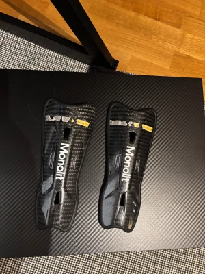 Monolit benskydd i kolfiber - Säljer ett par svarta benskydd från Monolit i kolfiber med vit logga. De har en smal och ergonomisk form med ventilationshål för extra komfort. Perfekt för dig som spelar fotboll och vill ha lätt och slitstark skyddsutrustning.