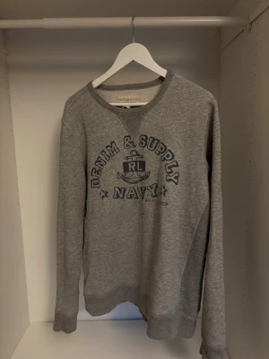 Grå tröja från Ralph Lauren - Riktigt fet grå sweatshirt från Ralph Lauren Denim & Supply med mörkblå tryck framtill. Riktigt skön tröja med snyggt tryck i riktigt bra skick.