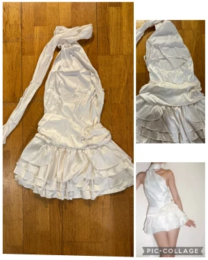 Vit satin kortklänning med volanger S - Supersnygg vit kortklänning i satin med omlott-design och oneshoulder. Klänningen har flera lager volanger i kjolen och en lång scarf-detalj som kan knytas runt halsen. Passformen är slim och den har en dekorativ blomdetalj i midjan. Perfekt för dig som vill sticka ut!